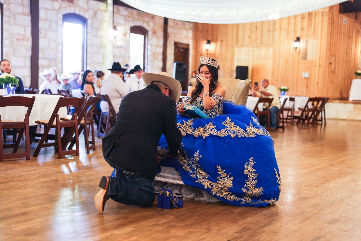 A young woman in a blue and gold quinceañera gown receives a crown from a man in a cowboy hat at a rustic venue.