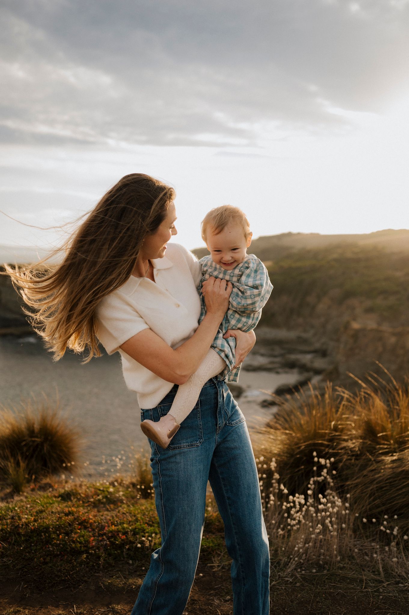 A mother holds her smiling baby outdoors at golden hour, her hair blowing in the wind near a coastal cliff.