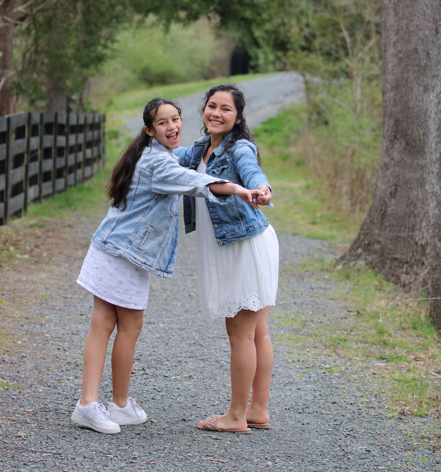 Two young girls in dresses and denim jackets laugh and pose together on a scenic gravel path outdoors.