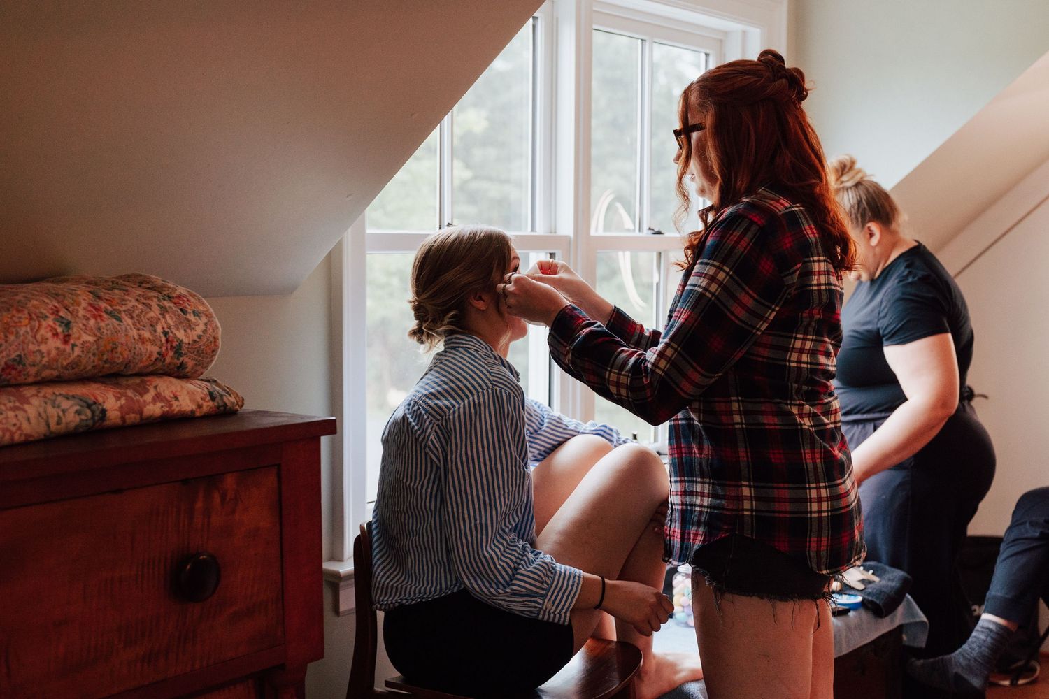 A woman applies makeup to a seated person near a window in a cozy attic room with another person watching.