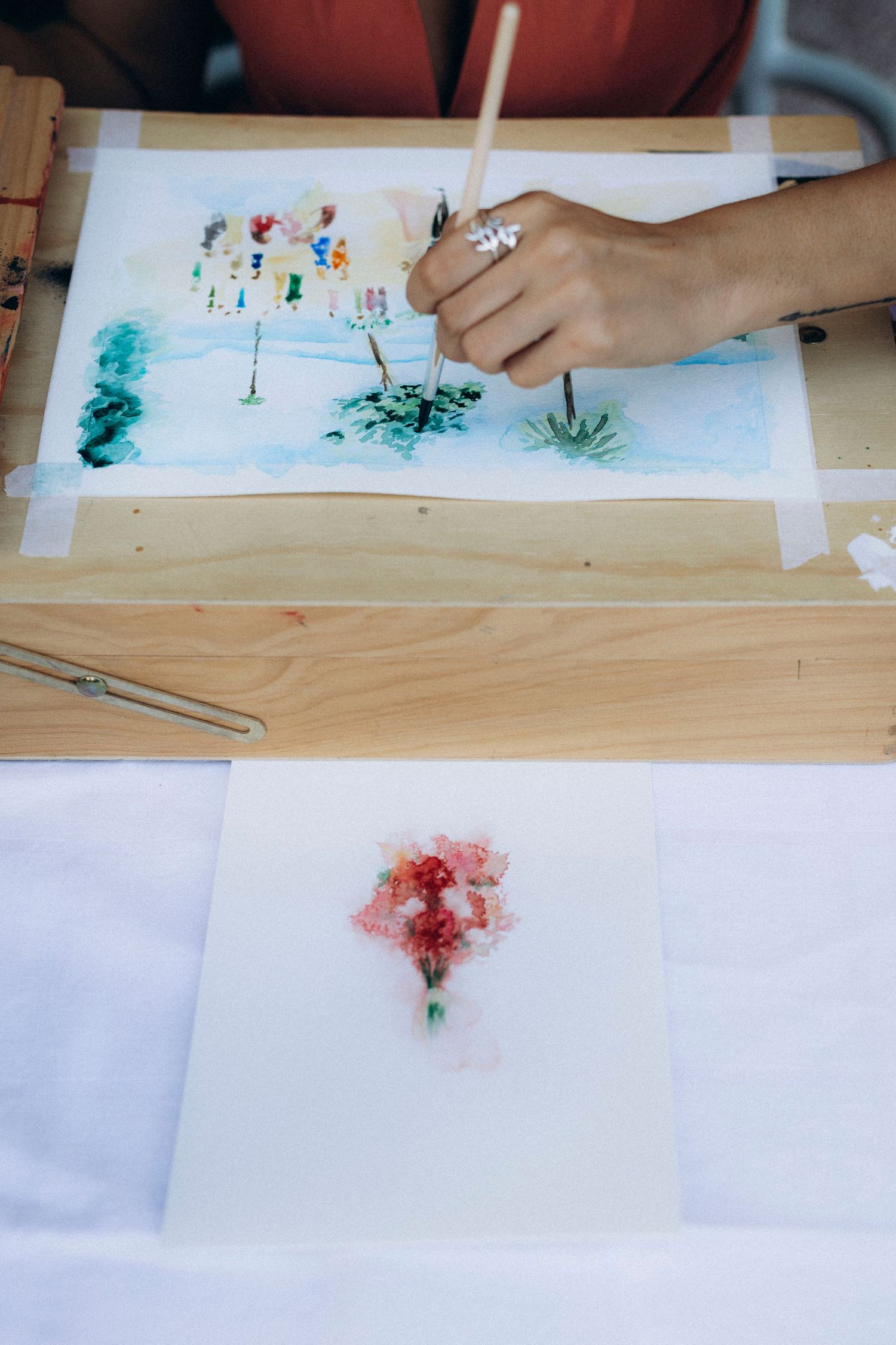 A tattooed woman artist in an orange dress holds a paintbrush at an outdoor table with string lights in the background.