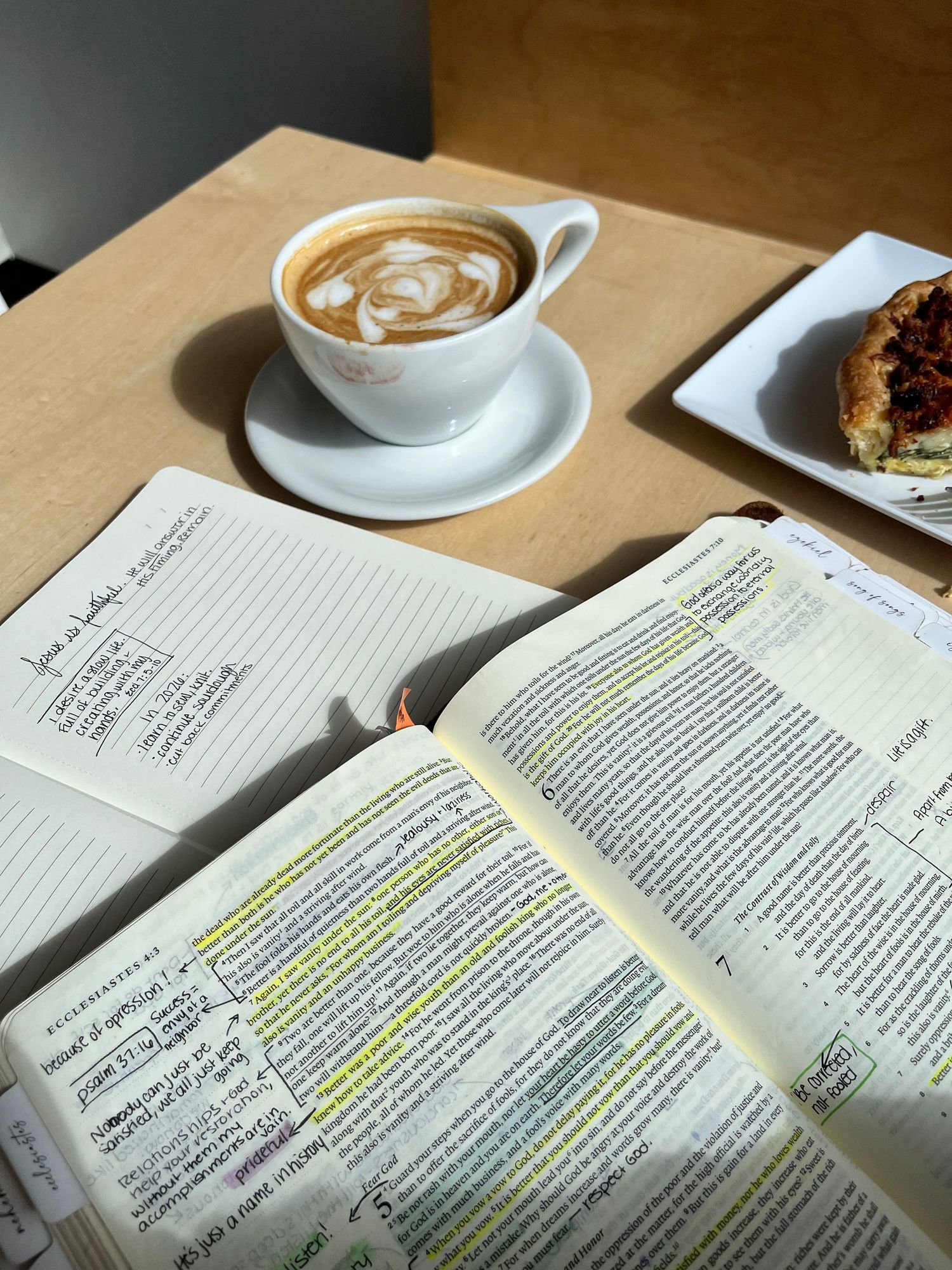 Open annotated book with highlighted text beside a latte art coffee cup and pastry on a wooden table.