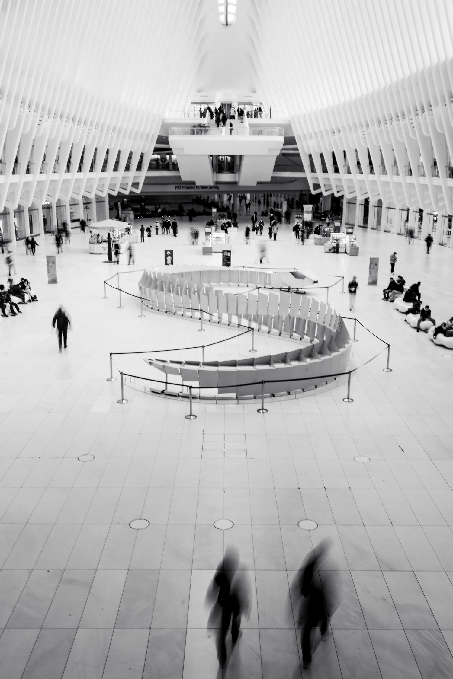 Scatto in bianco e nero all'interno dell'Oculus con un'installazione artistica sinuosa e sagome di persone in movimento mosso.