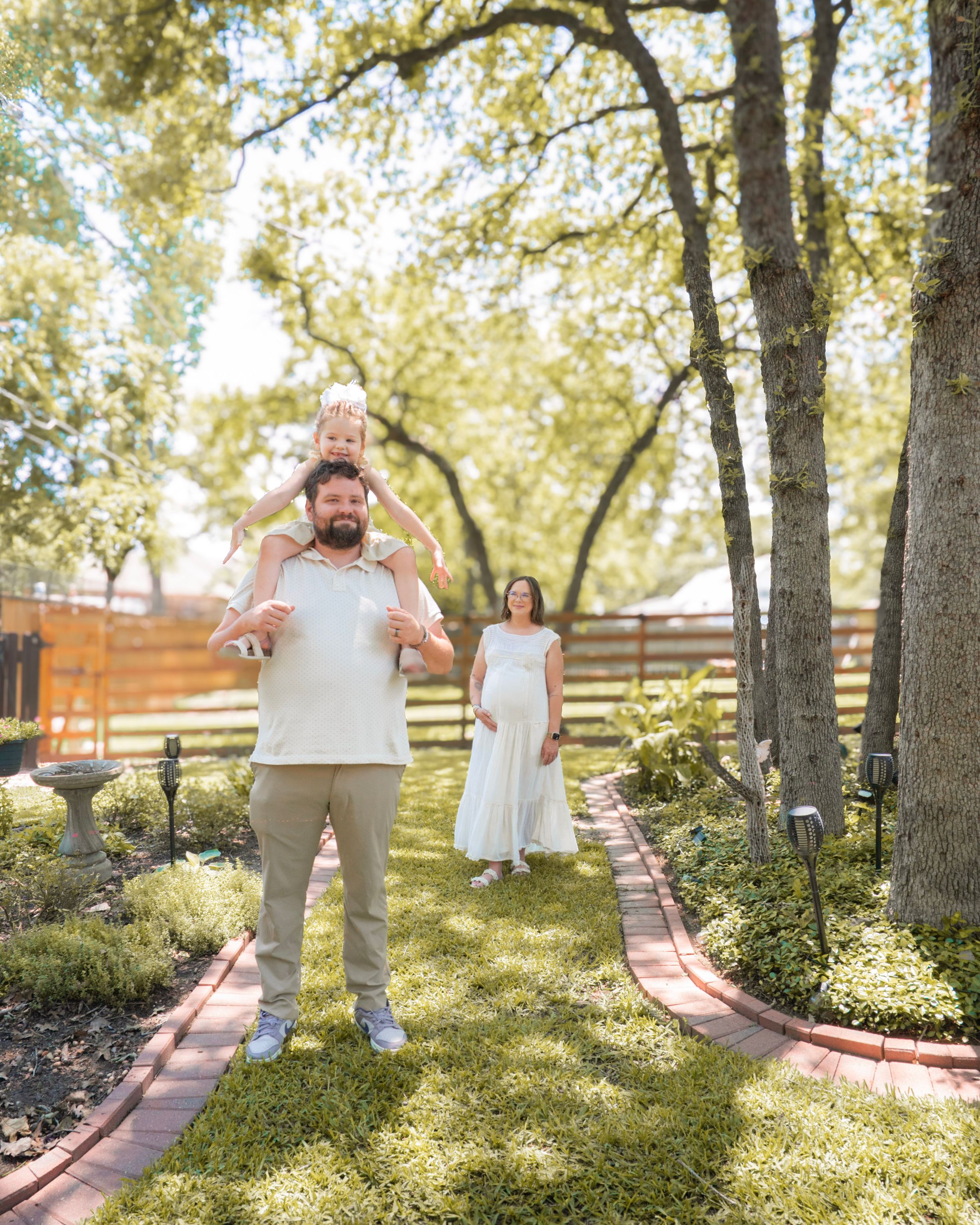 Father carries toddler on shoulders while pregnant mother walks behind in a lush garden path.