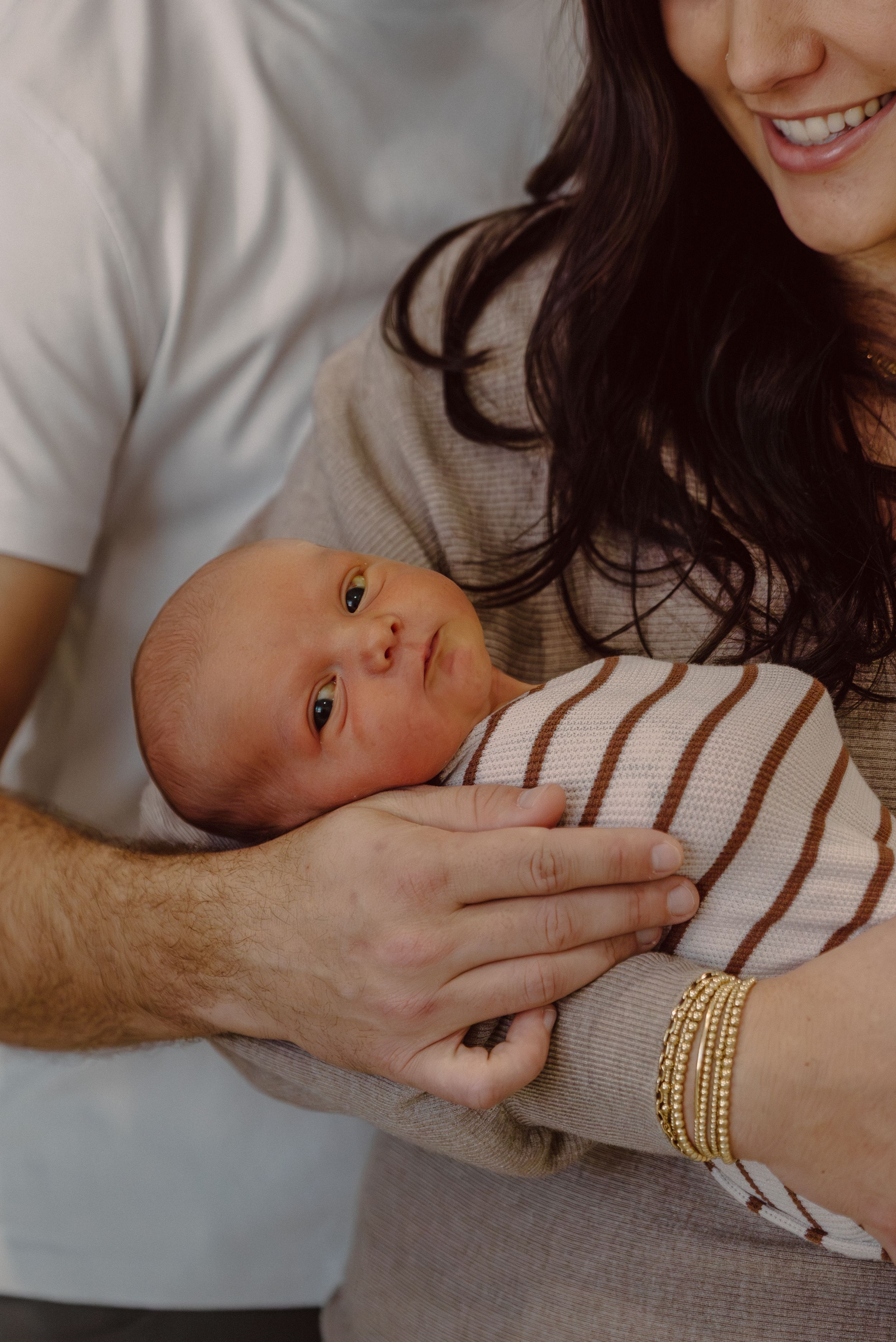 New parents smiling as they cradle their newborn baby wrapped in a striped swaddle blanket.