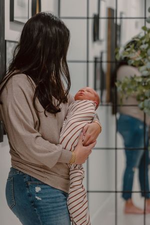 A dark-haired woman holds a swaddled newborn baby in a striped blanket indoors.