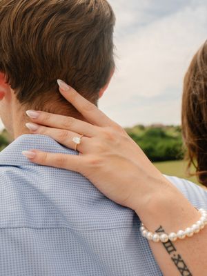 Woman rests hand on mans shoulder displaying a stunning diamond engagement ring and pearl bracelet.