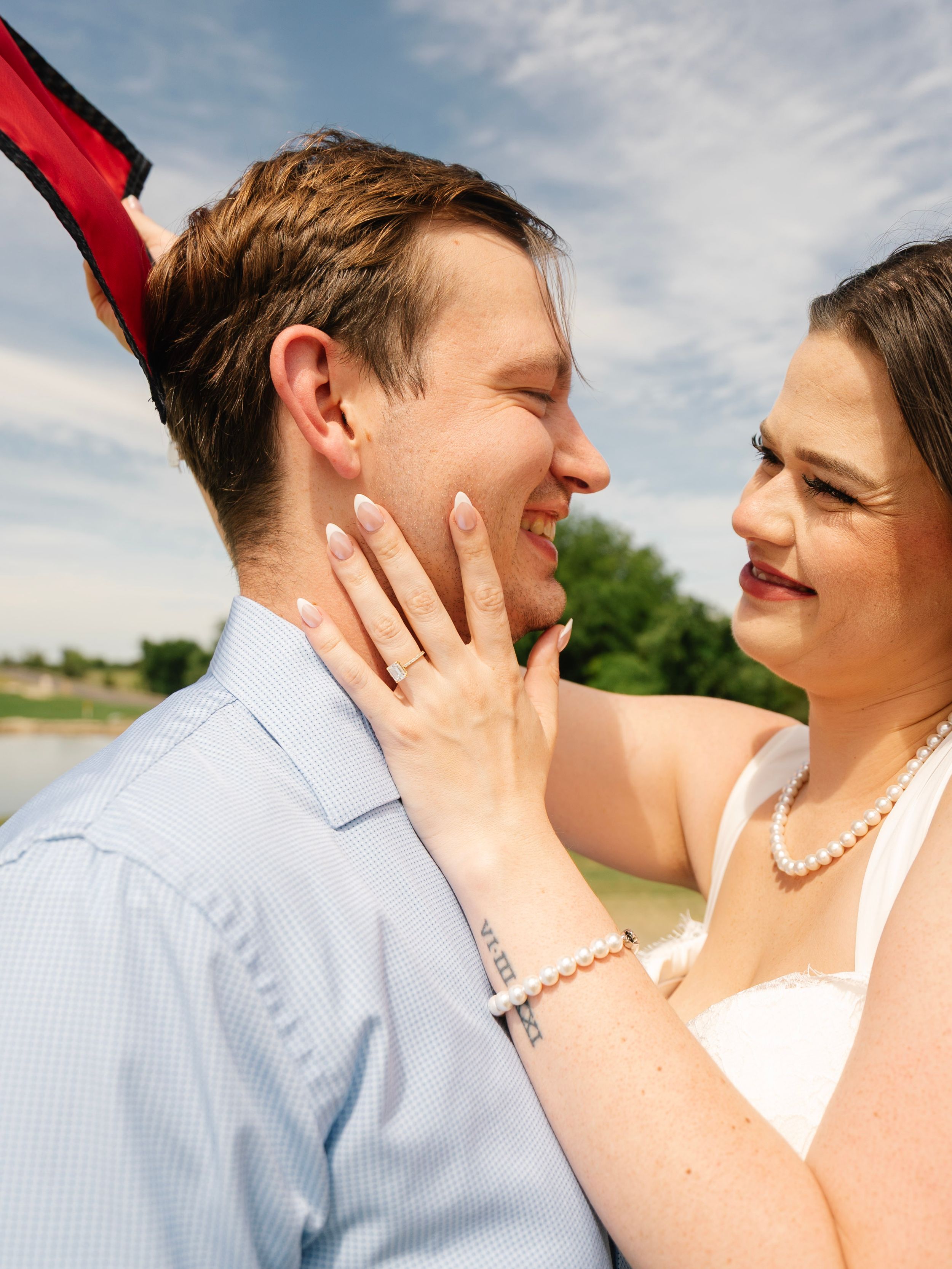 Smiling couple in outdoor engagement photo, woman showing diamond ring on man's face.