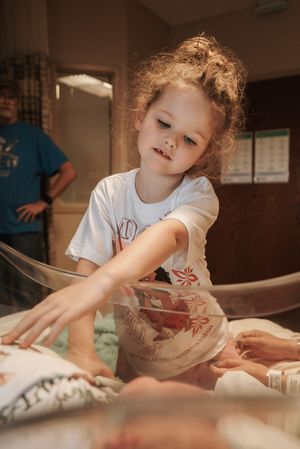 A young girl with curly hair reaches into a hospital bassinet to touch a newborn baby.