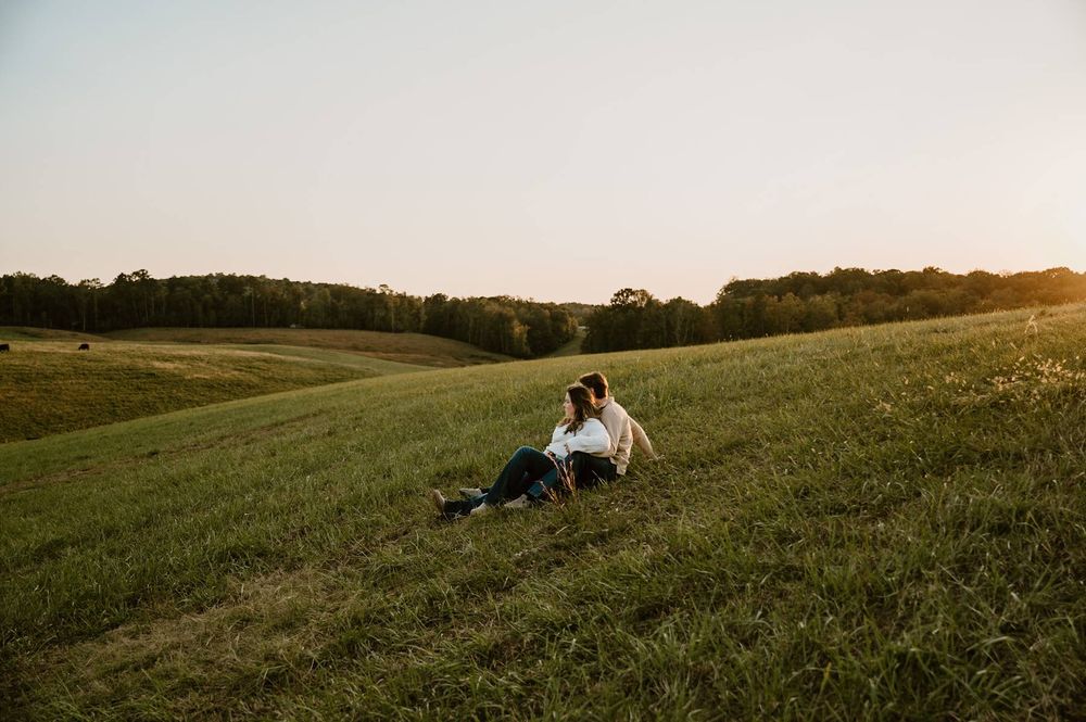 Couple sitting together on a lush green hillside field at golden hour sunset engagement session, Lexington NC