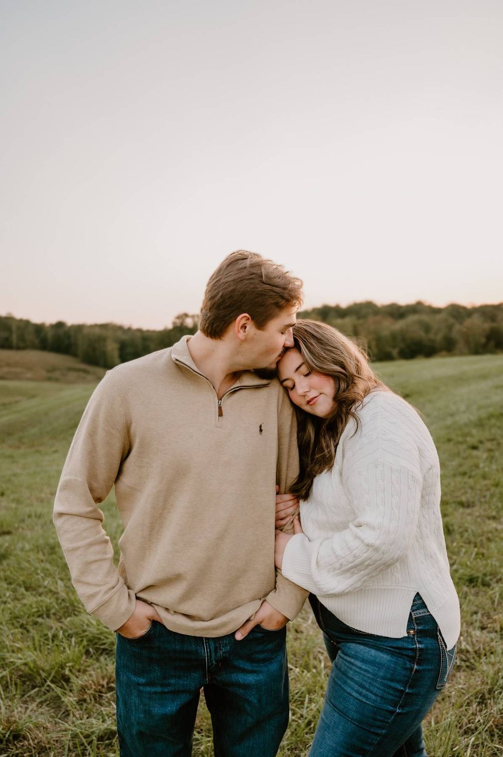 Man in beige pullover kissing woman on forehead during golden hour field engagement photo session in High Point NC