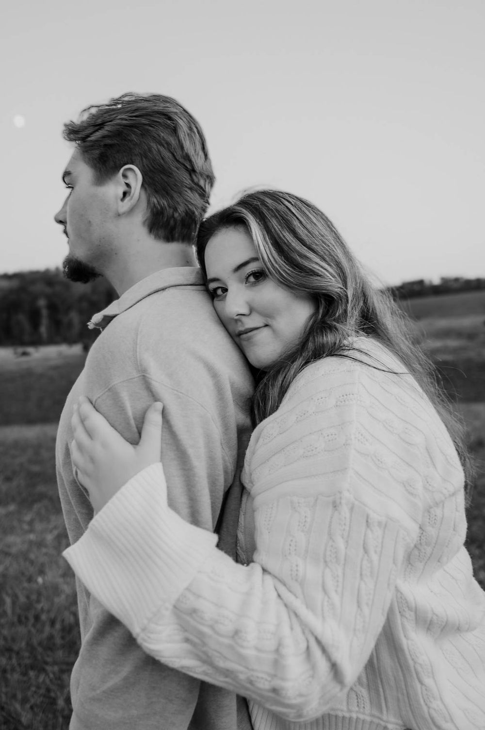 Black and white photo of woman hugging man from behind in an open field at dusk, couples photographer Greensboro NC
