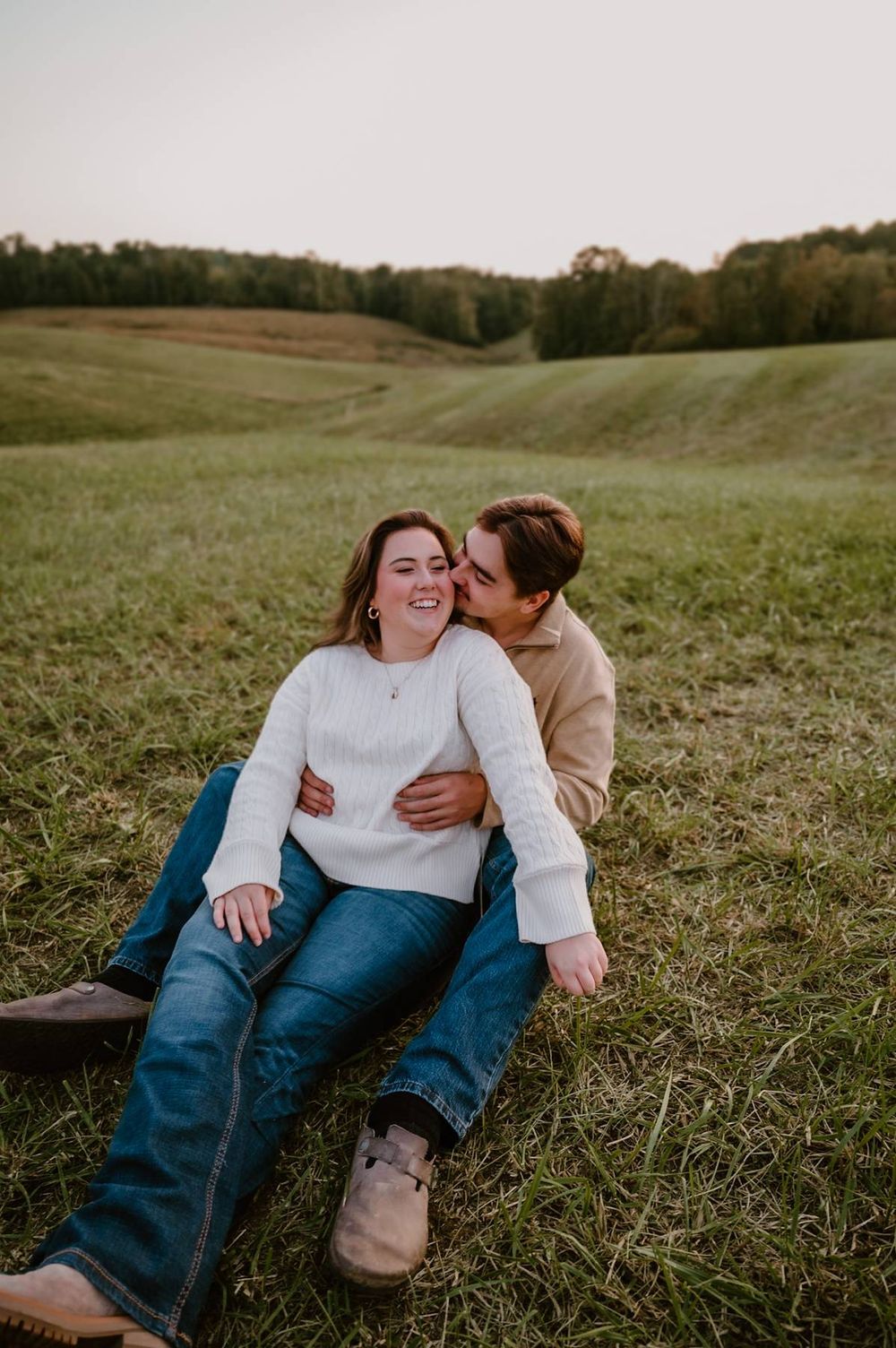 Couple sitting in green field, man kissing woman's cheek as she laughs, wearing white sweater and jeans, couples photographer in North Carol