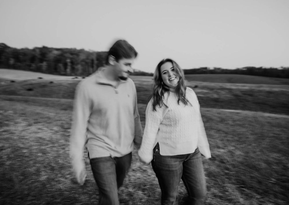 Black and white photo of couple holding hands and laughing while walking through an open field, couples photographer in North Carolina