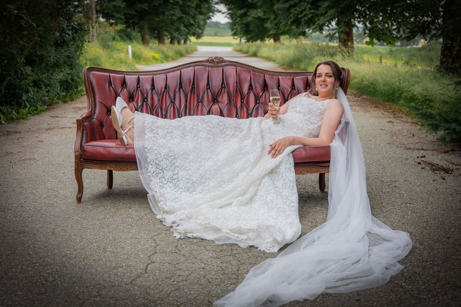 Bride in lace wedding gown reclines on vintage red sofa holding champagne glass on scenic road.