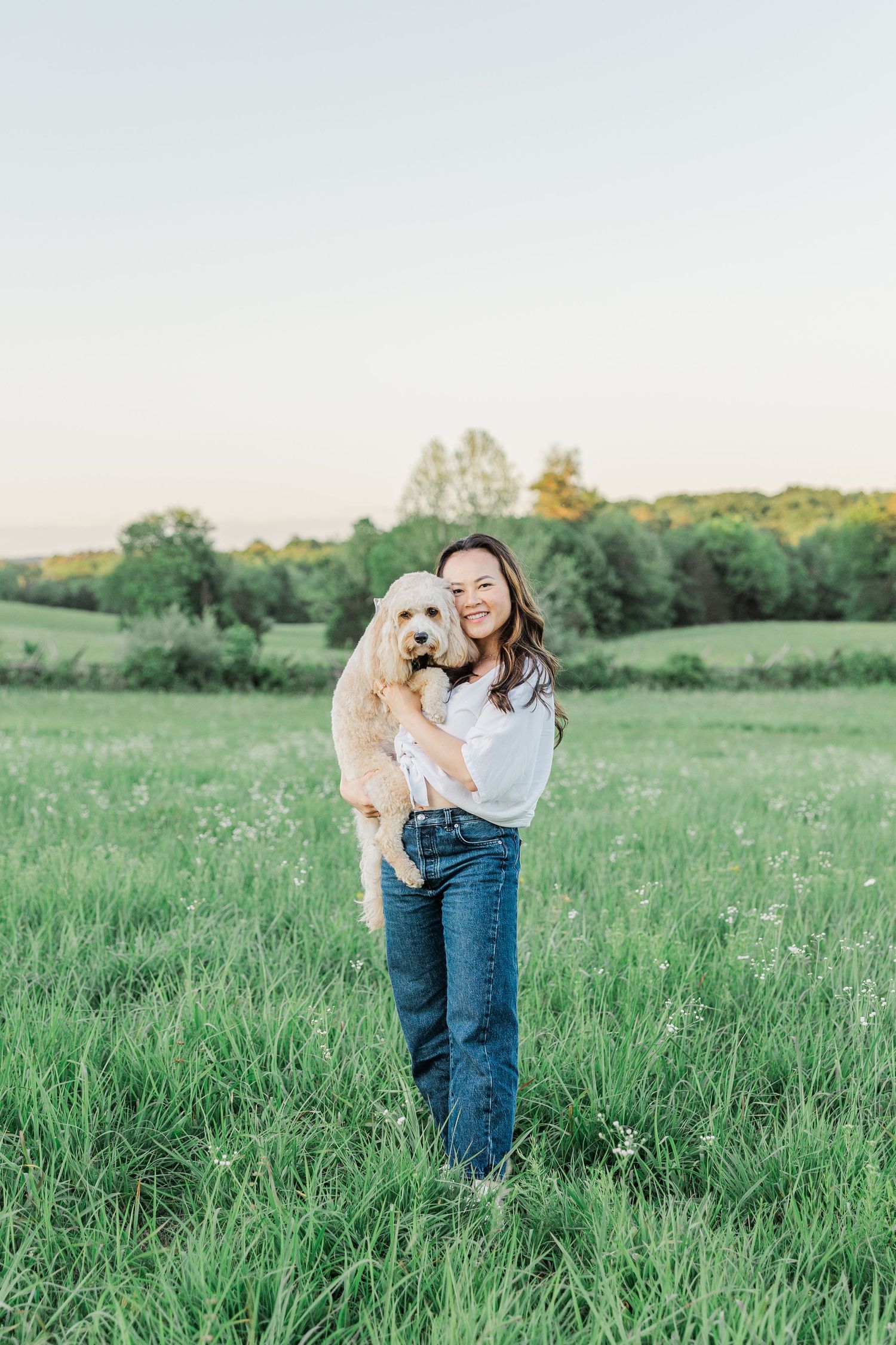 A smiling woman holds a fluffy golden doodle dog while standing in a lush green field at sunset.