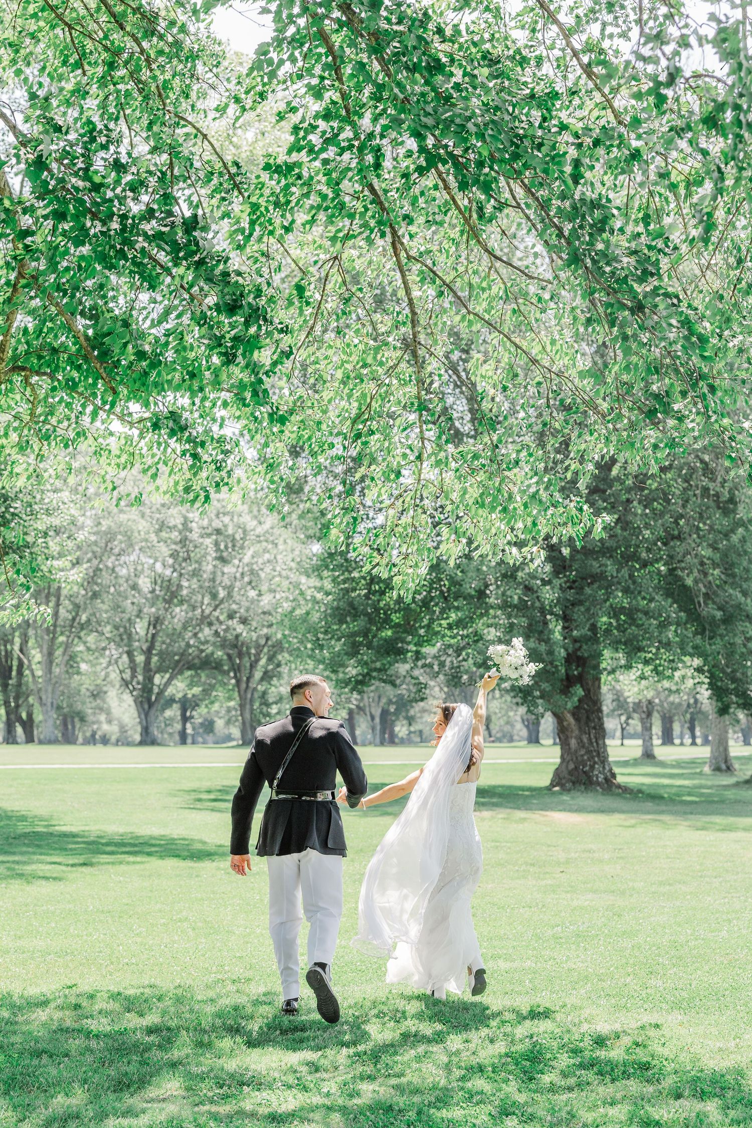 Bride and groom walking hand in hand through a lush green park, bride holding bouquet aloft in her wedding dress.