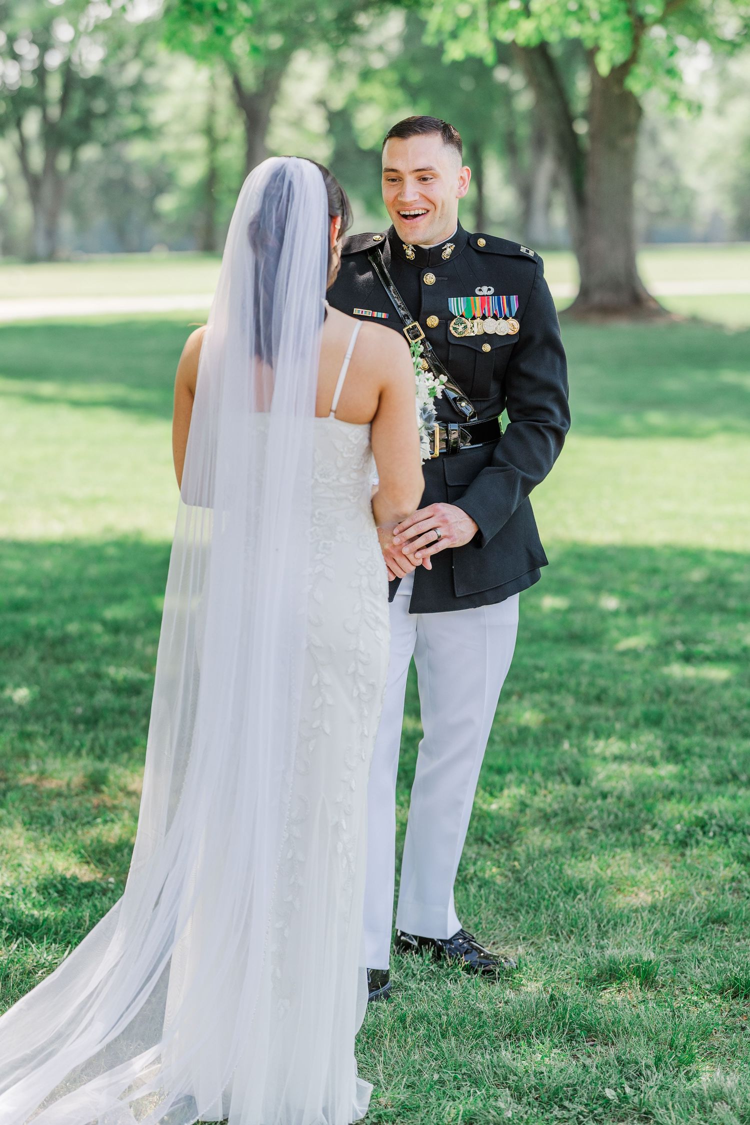 Military groom in dress uniform laughing with bride in lace wedding gown and veil during outdoor first look.