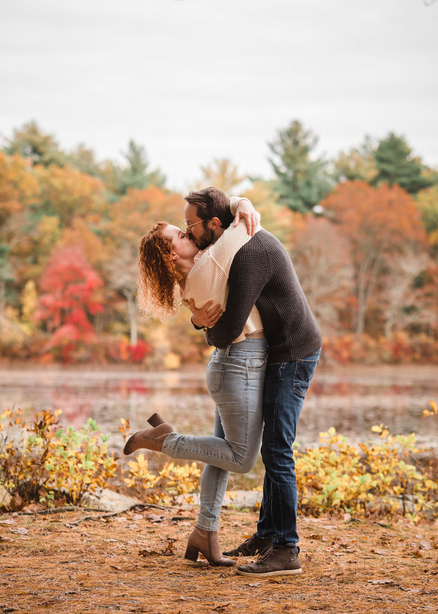 Engaged couple kissing at Lake Leach in Borderland State Park, her foot kicked up, autumn foliage reflected in the water.
