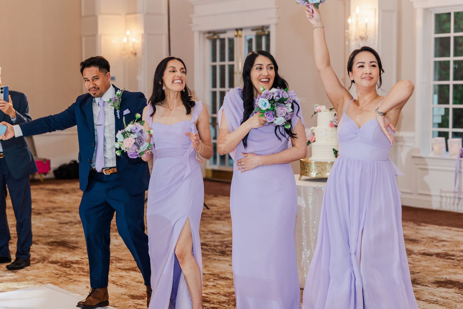 Bridesmaids in lavender gowns celebrate joyfully at a wedding reception with a white cake in the background.