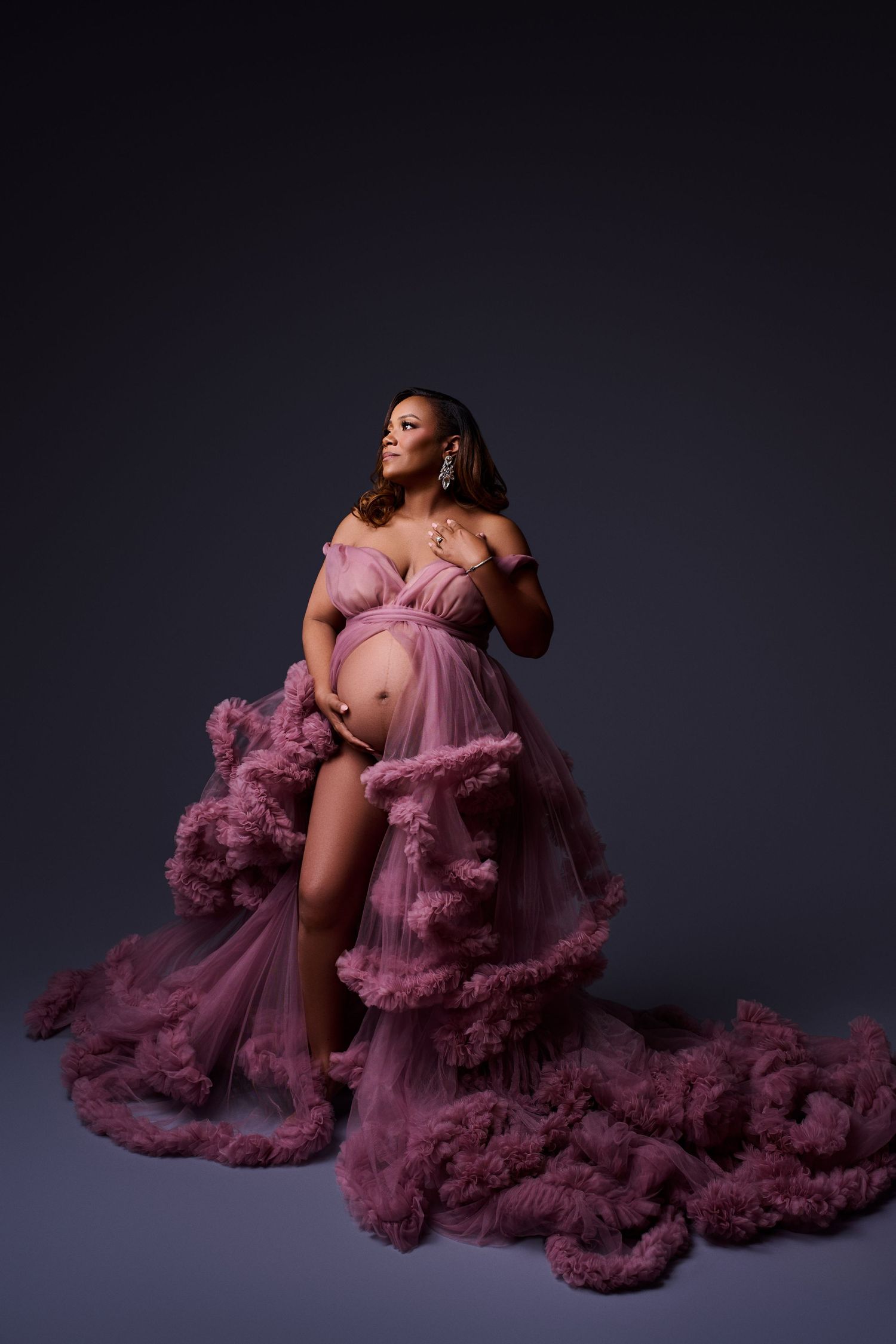 Pregnant woman in flowing pink tulle maternity gown posing elegantly in a professional studio photo shoot.