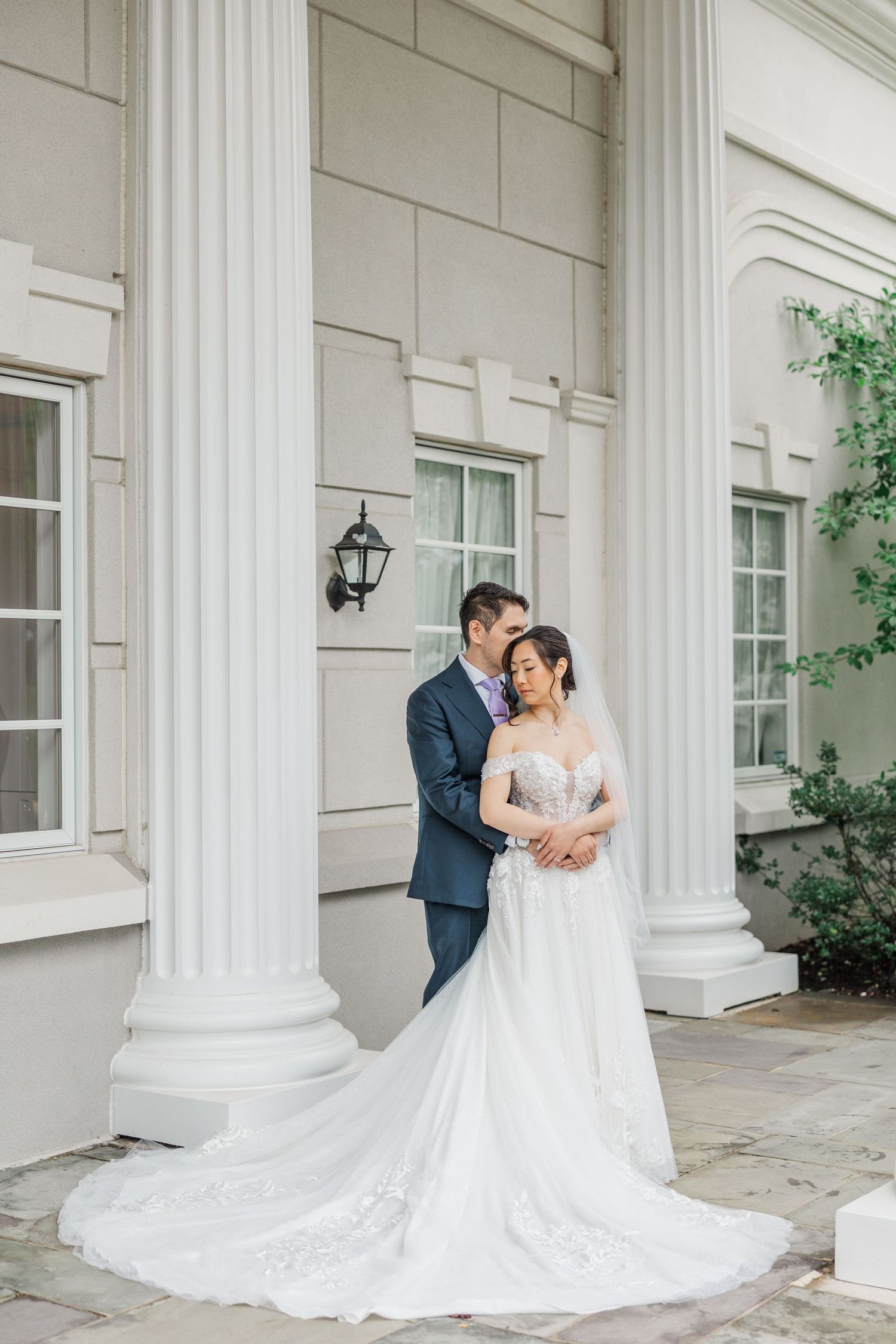 Bride in lace gown and groom in navy suit pose romantically between white columns of elegant venue.