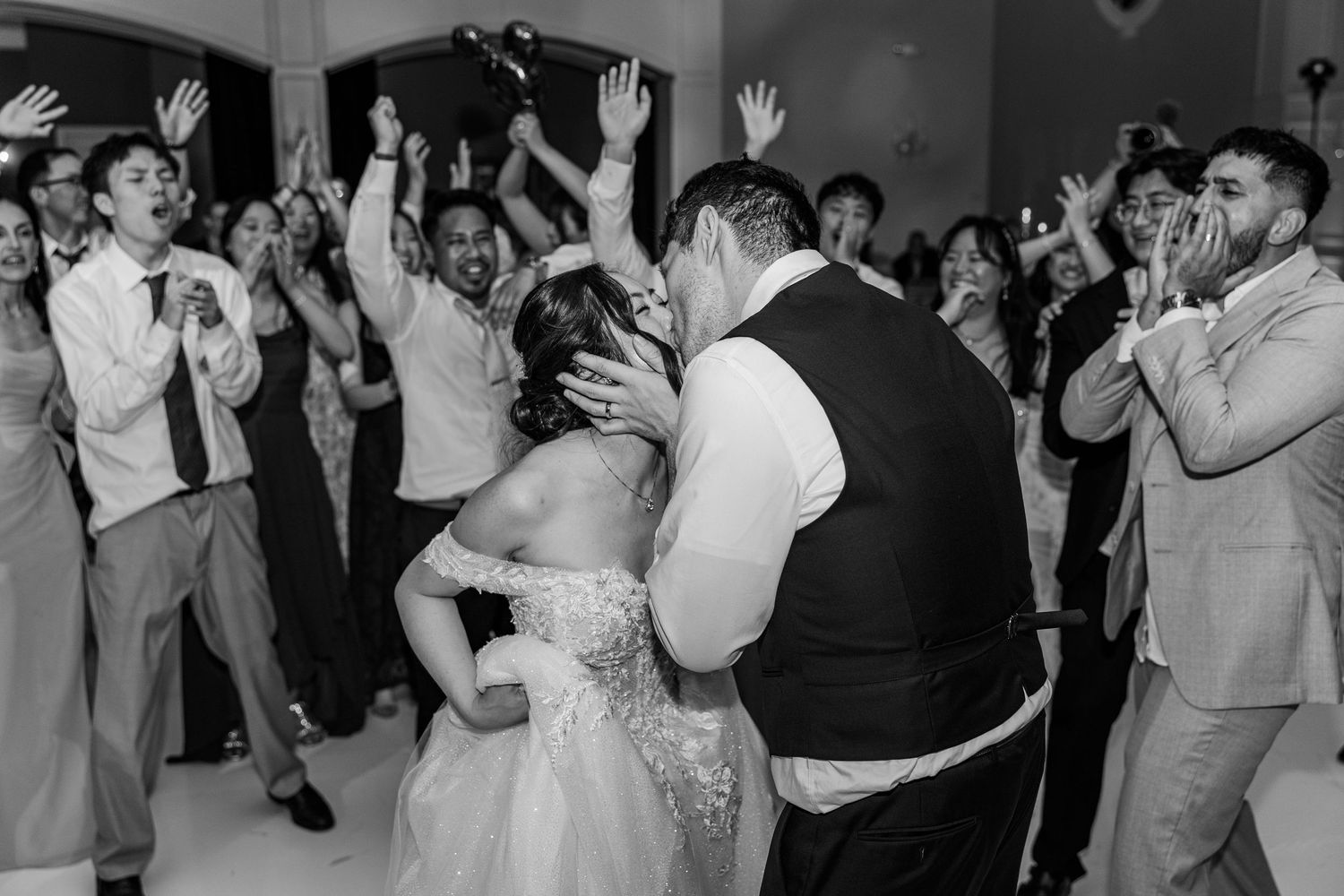 Black and white photo of bride and groom sharing a kiss on the dance floor surrounded by cheering wedding guests.