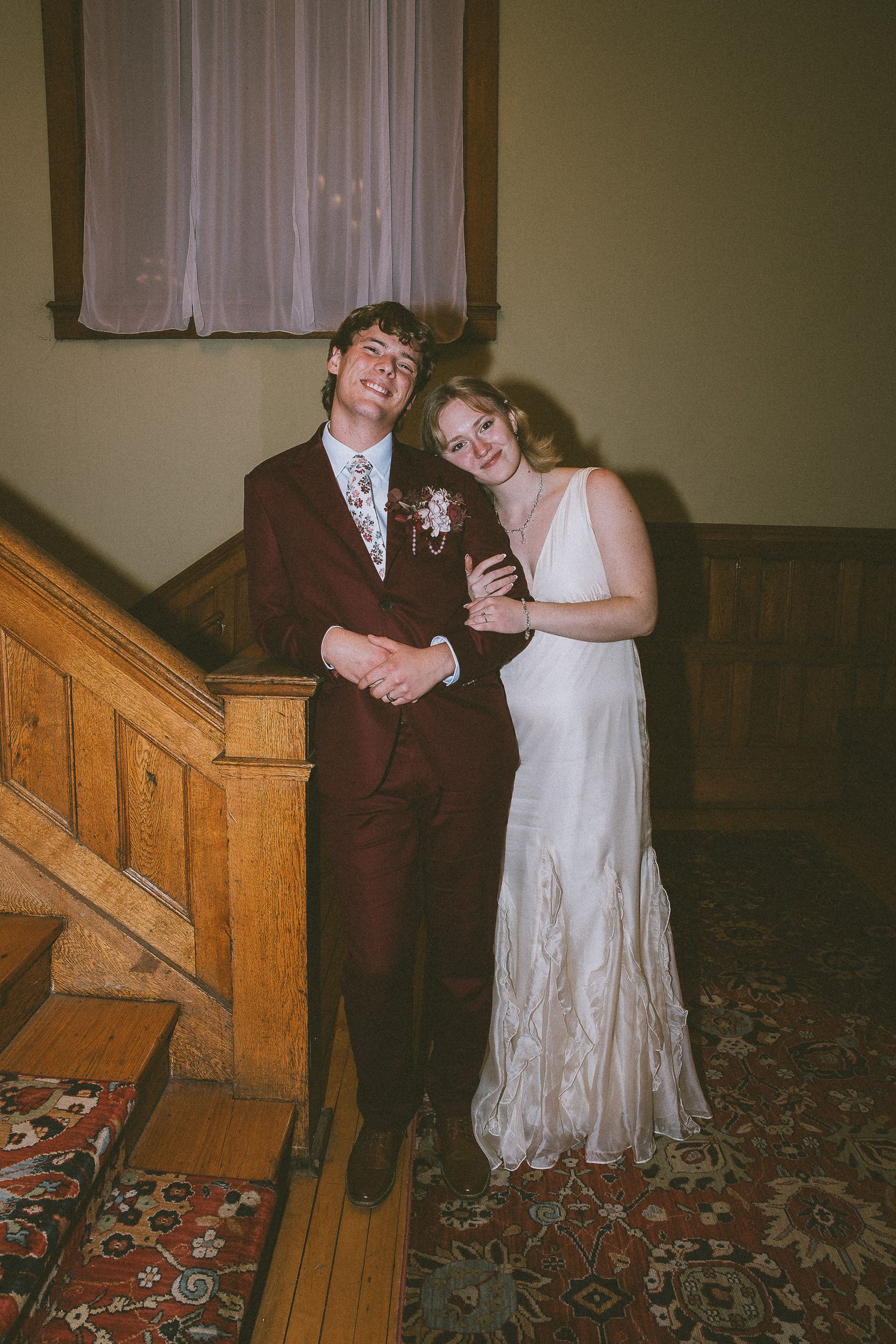 A smiling couple in wedding attire pose by a wooden staircase, groom in burgundy suit, bride in white gown.