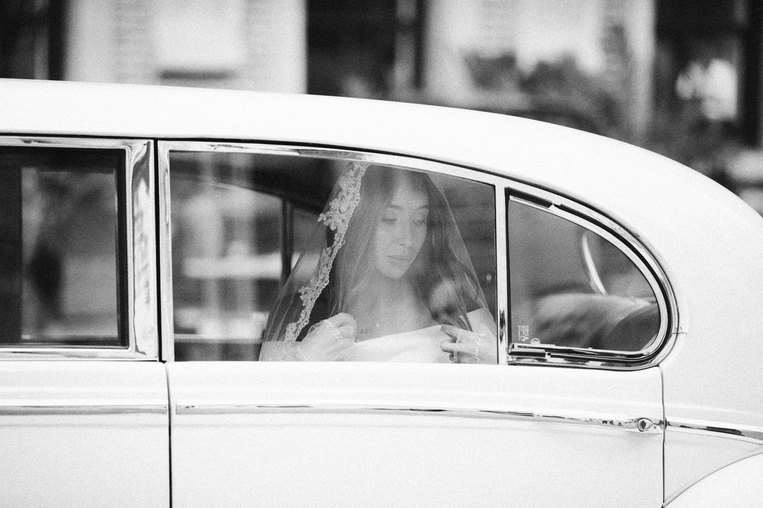 A bride in a veil sits inside a vintage white car, captured in elegant black and white photography.