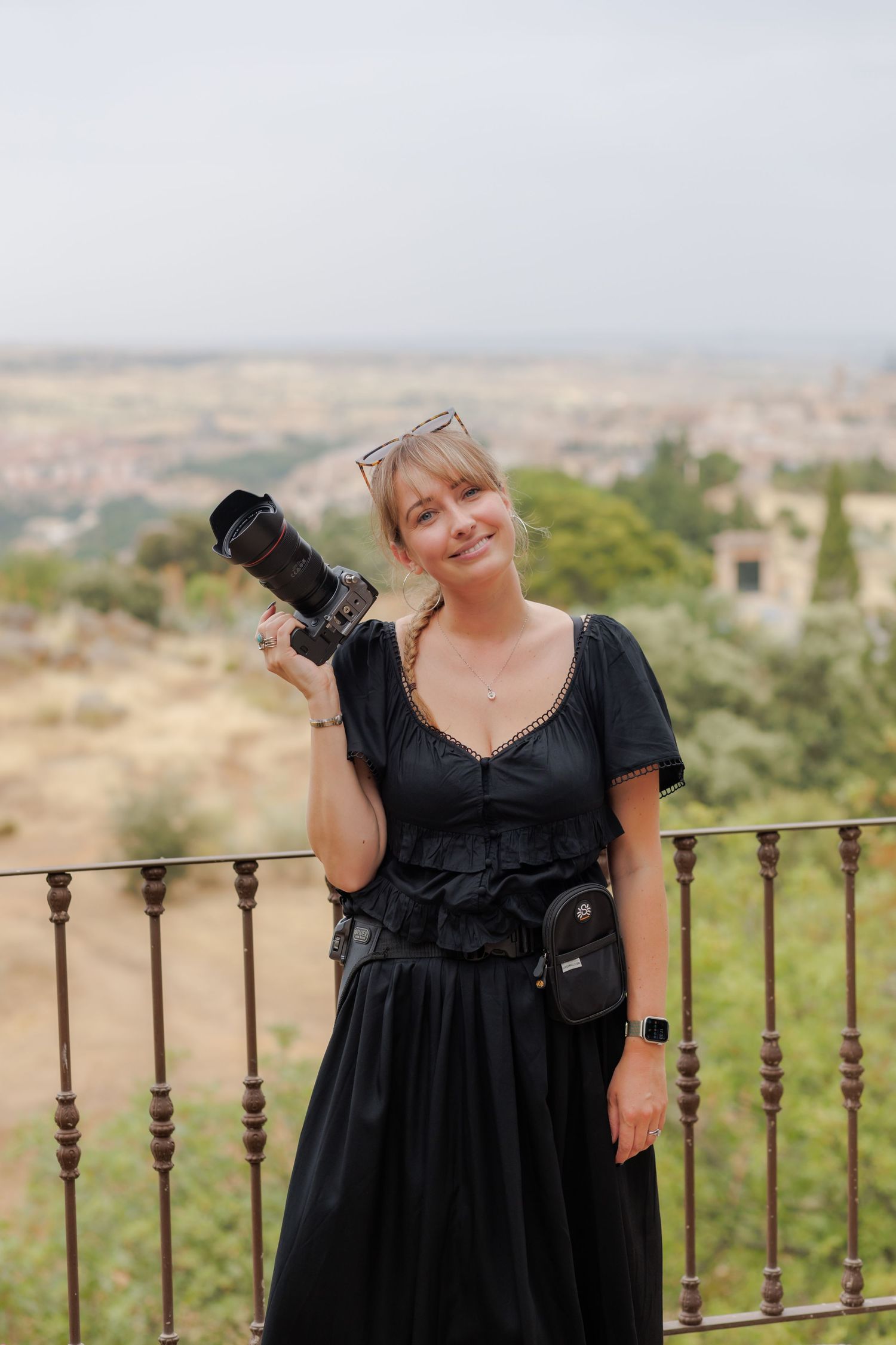 Woman in black dress holding a camera on a balcony with scenic landscape views in the background.