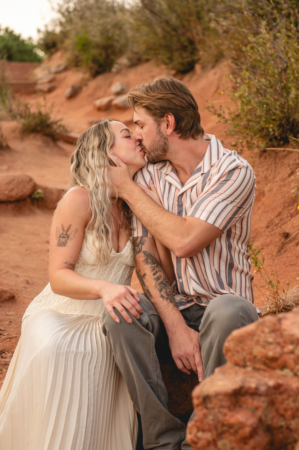 Couple sharing a romantic kiss on red rock terrain, woman in white dress with tattoos, man in striped shirt.