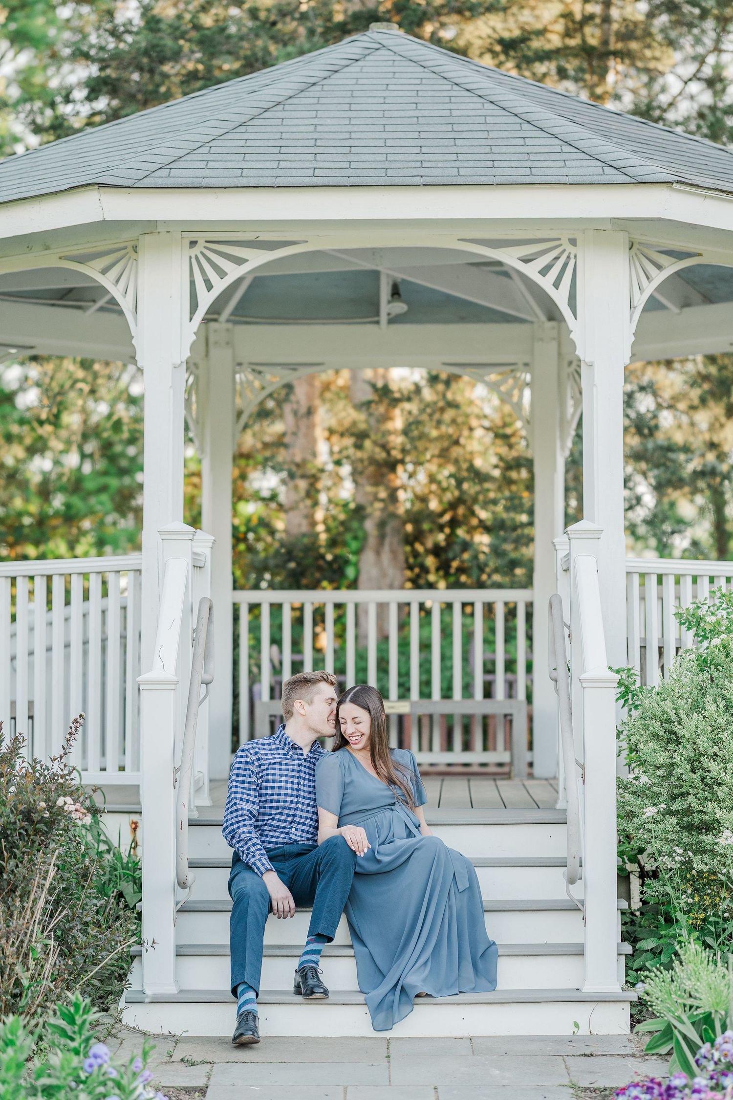 A couple sits together on gazebo steps in a lush garden, the woman in a flowing blue gown cradling her baby bump.