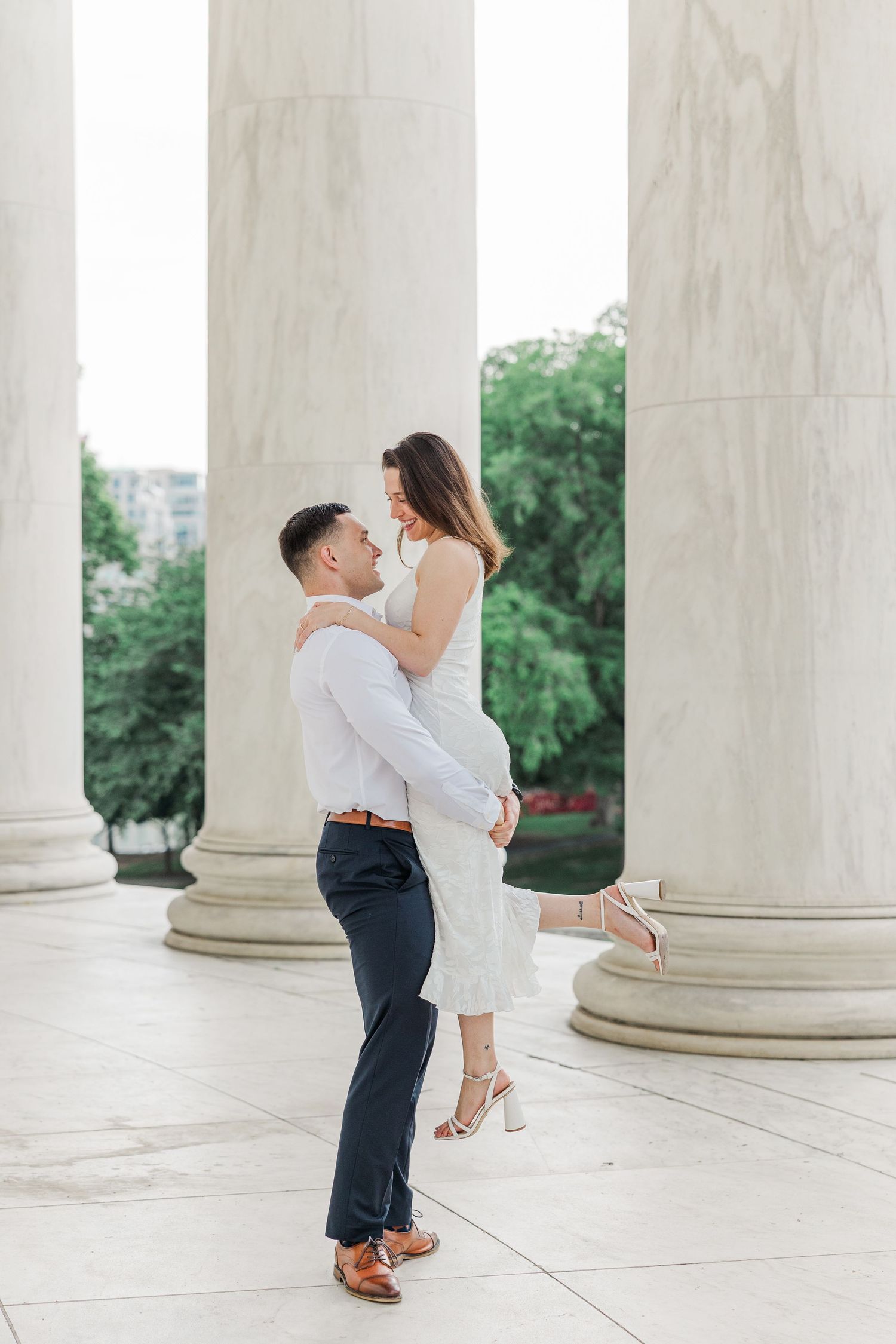 A couple shares a romantic moment among white marble columns, as a man lifts a woman in a white dress at the Jefferson Memorial.