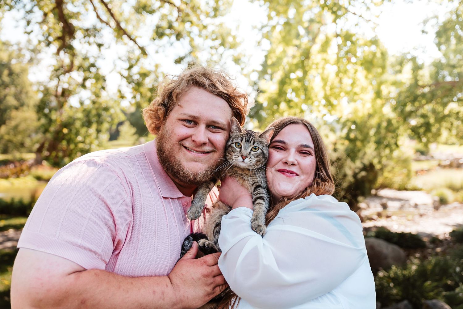 Couple smiling outdoors holding a tabby cat, surrounded by lush green trees in a sunny park setting.