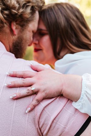 Couple embracing closely, woman wearing engagement ring with manicured nails, man in pink sweater, romantic outdoor setting.