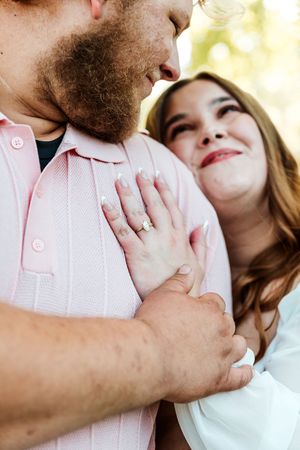 Couple embracing outdoors, woman smiling up at bearded man, engagement ring visible on her hand resting on his pink shirt.