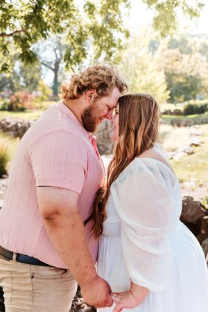 A couple in a sunny park shares a tender forehead touch, the woman in white and man in pink holding hands.