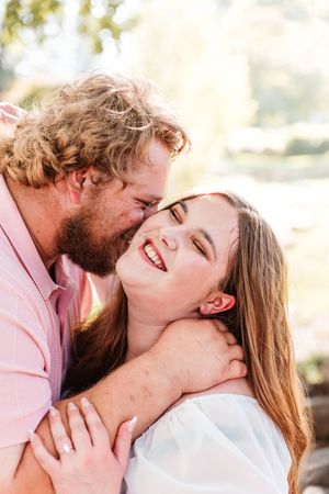 A curly-haired bearded man kisses a laughing brunette woman outdoors in warm sunlight.