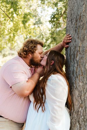 A couple shares a romantic kiss leaning against a tree outdoors, man in pink shirt, woman in white dress.