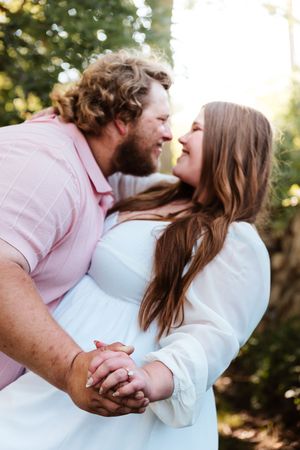A couple holds hands and gazes lovingly at each other outdoors, the woman in white dress and man in pink shirt.