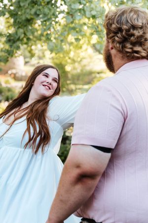 A smiling woman in a light blue dress holds hands with a curly-haired man in a pink shirt outdoors.