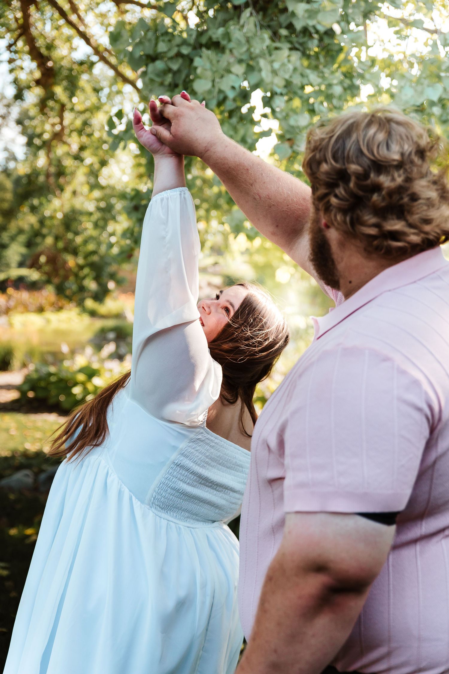 A couple dances outdoors, woman in white dress reaches up holding hands with curly-haired man in pink shirt amid green trees.