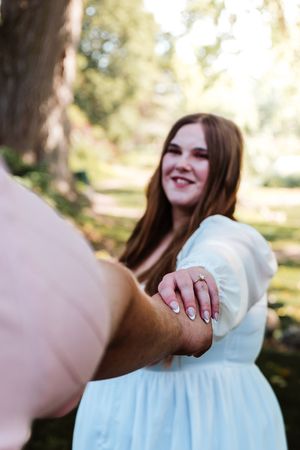 Smiling brunette woman in light blue dress showing engagement ring while holding hands outdoors in a sunny park.