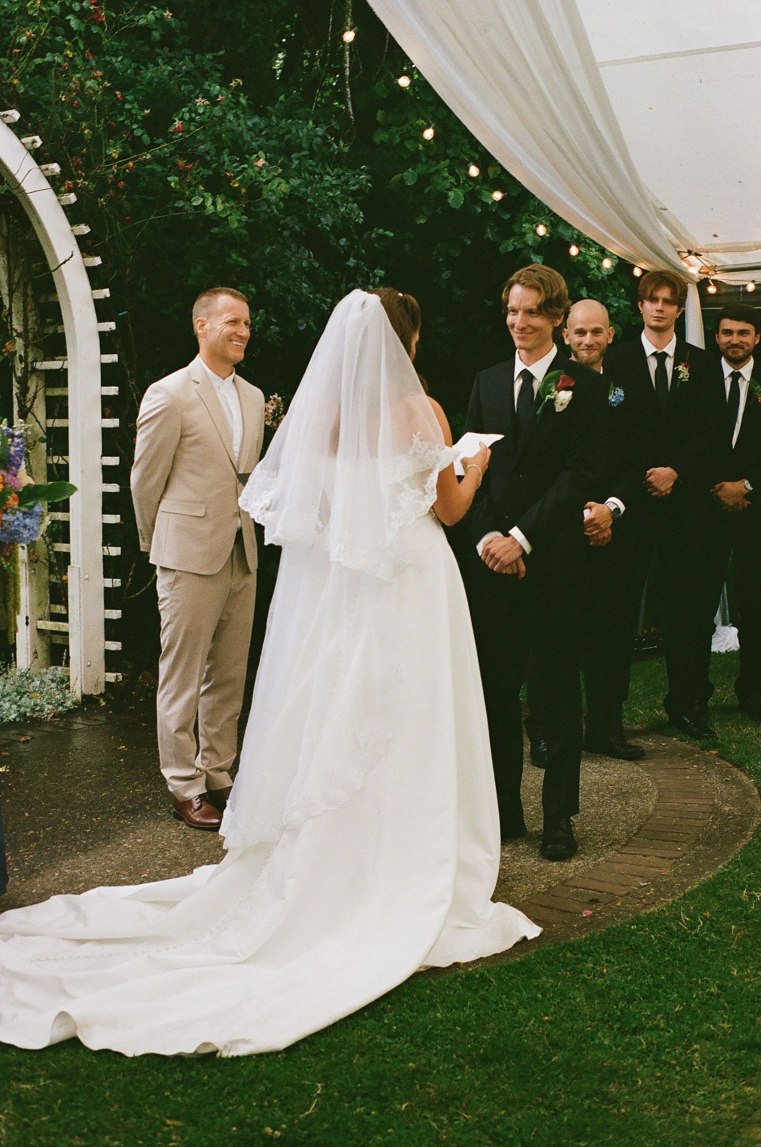Outdoor wedding ceremony with bride in white gown and veil exchanging vows with groom in black tuxedo, groomsmen standing behind.