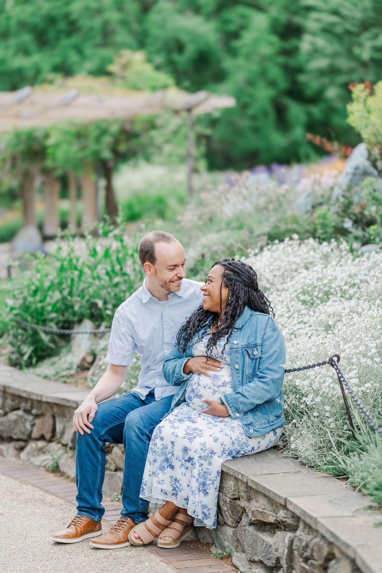 A couple sits on a stone wall in a lush garden, smiling at each other, the woman in a floral dress cradling her baby bump.