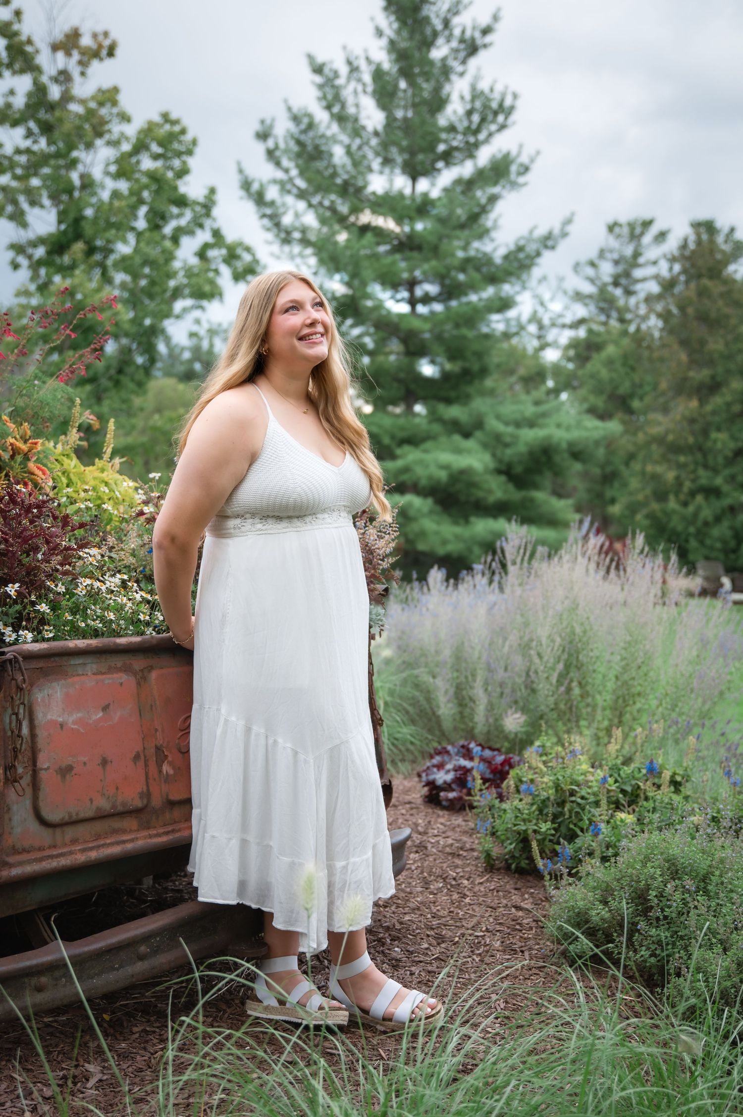 A smiling woman in a white sundress and sandals stands in a lush garden surrounded by wildflowers and evergreen trees.