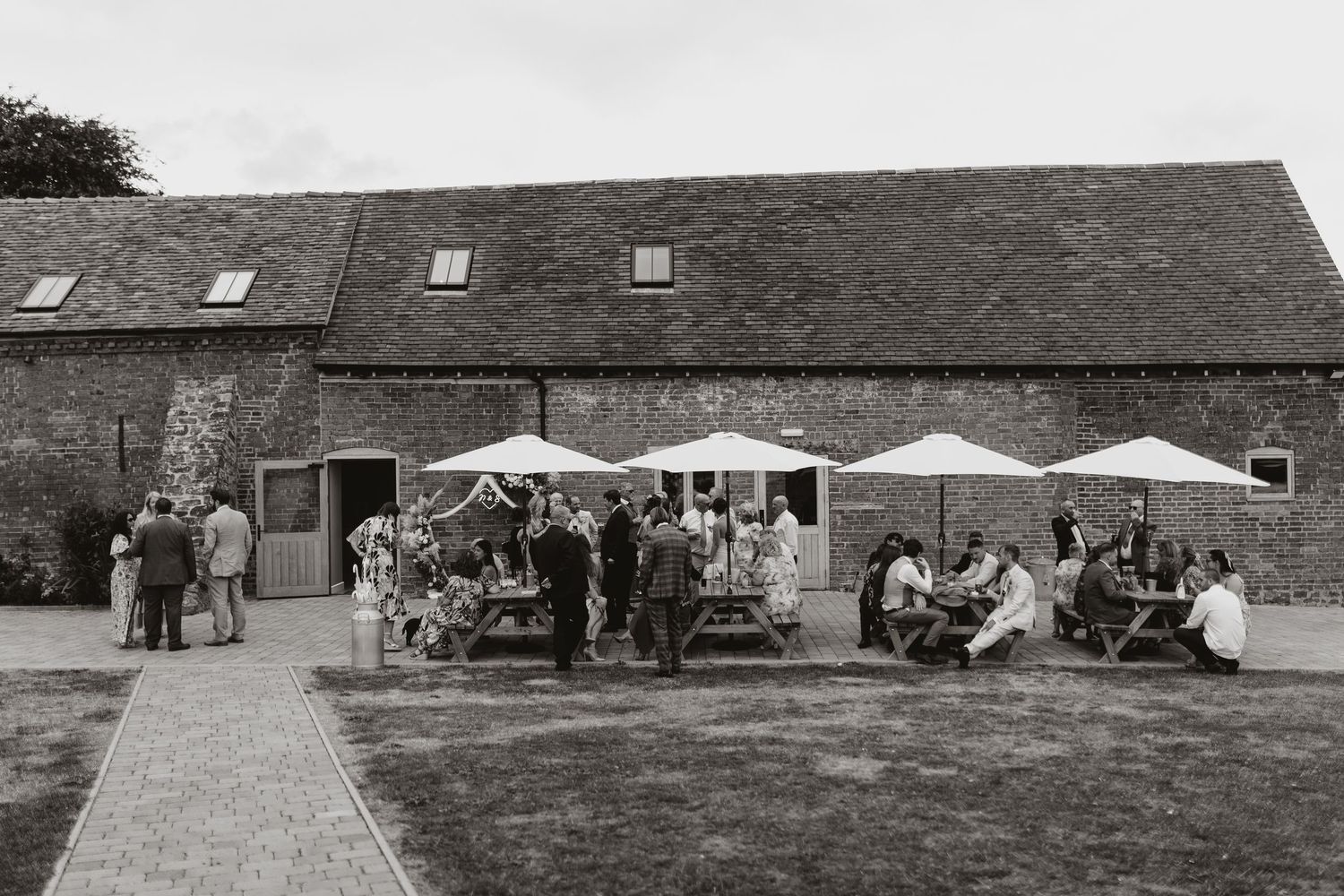 Black and white photo of guests socialising outside a rustic barn venue with picnic tables and white umbrellas at Moat Hall Barns