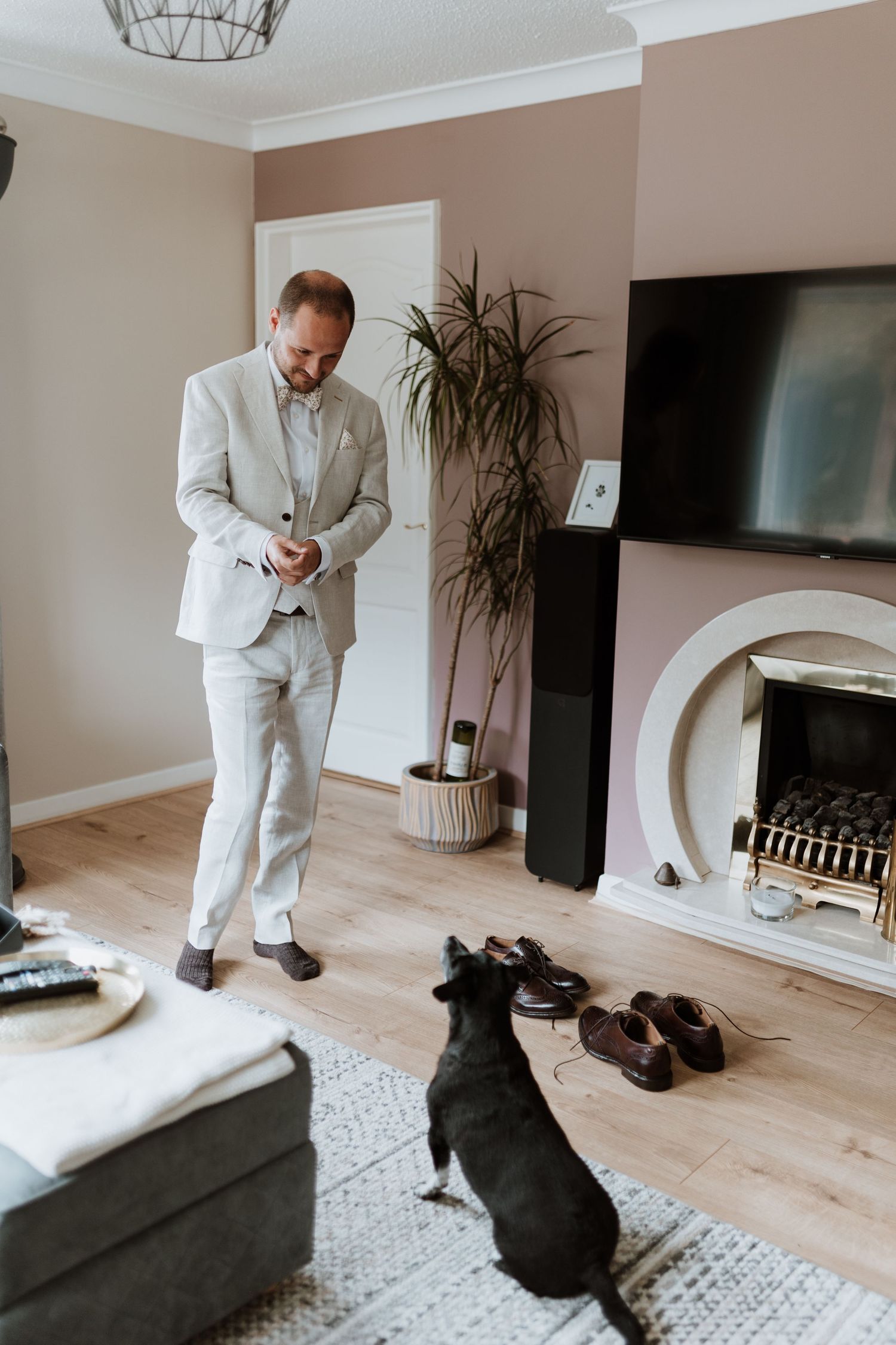 Groom in light grey suit stands in modern living room with dog, fireplace, and dress shoes on floor.