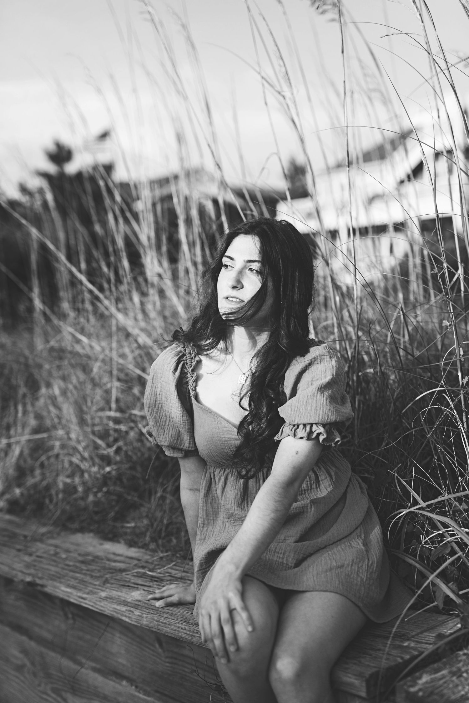 A person in a vintage dress sits on a wooden bench among tall grasses in black and white.