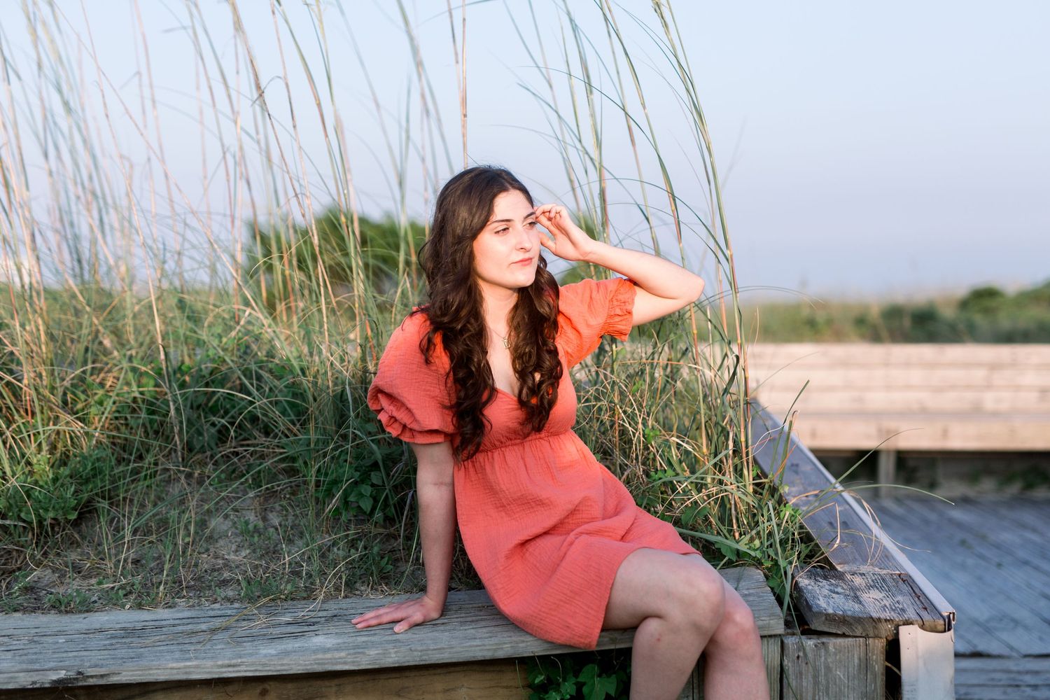 A person in a coral dress poses dreamily on a wooden deck surrounded by sea grass.
